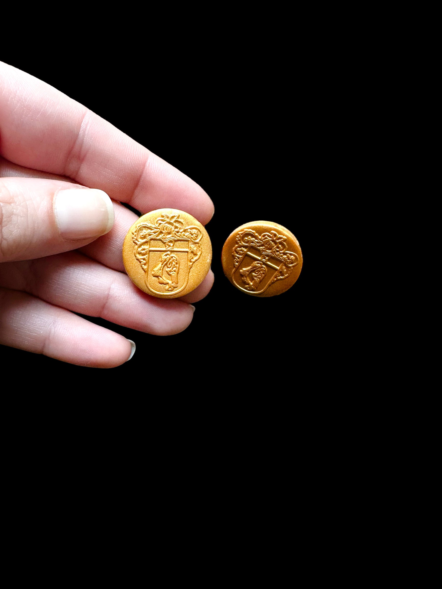 A hand holding gold handmade earrings. These hypoallergenic earrings are embossed with the FLORIT family crest, offering a touch of heritage and elegance to your outfit.