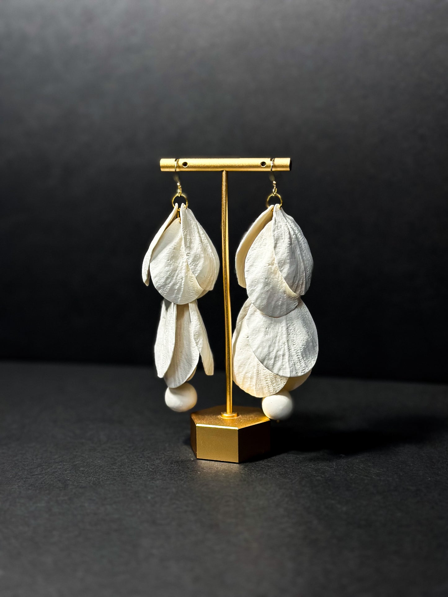 White handmade earrings hanging on an earring display. Meticulously hand sculpted petals are combined to achieve a cascading floral design.